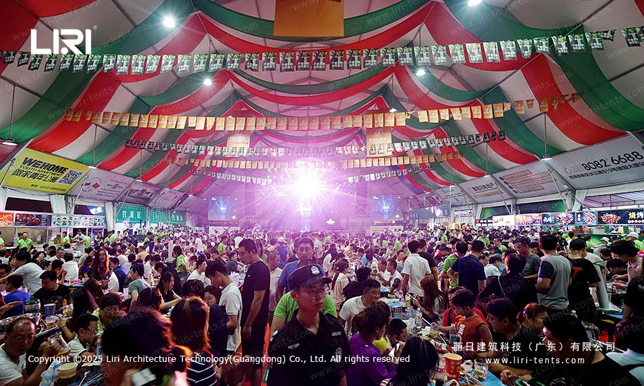 Crowded beer festival tent dining area with lighting and vendor booths